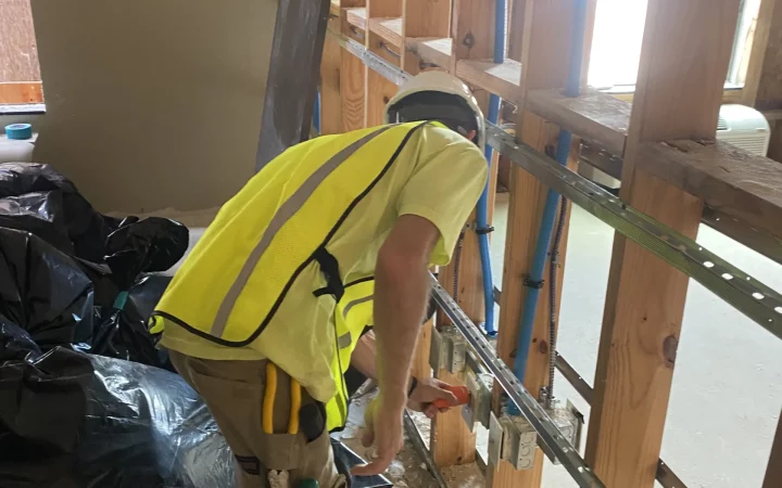 An electrician in safety gear installs electrical components within an open wall framework.