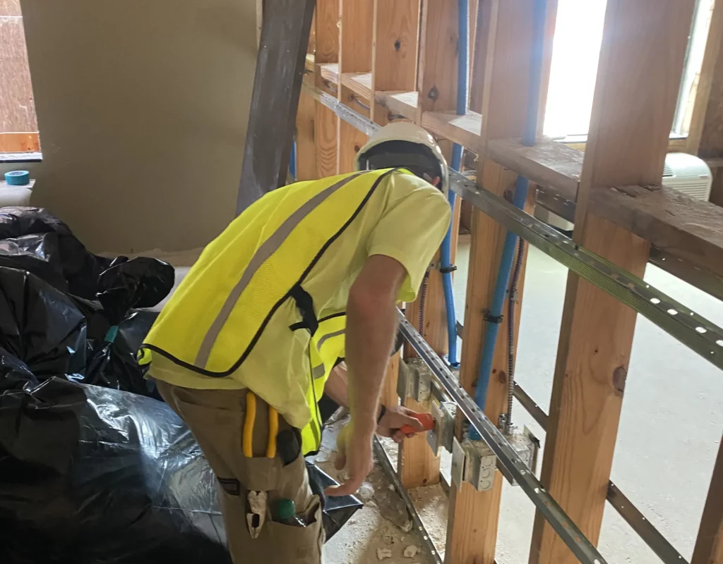 An electrician in safety gear installs electrical components within an open wall framework.