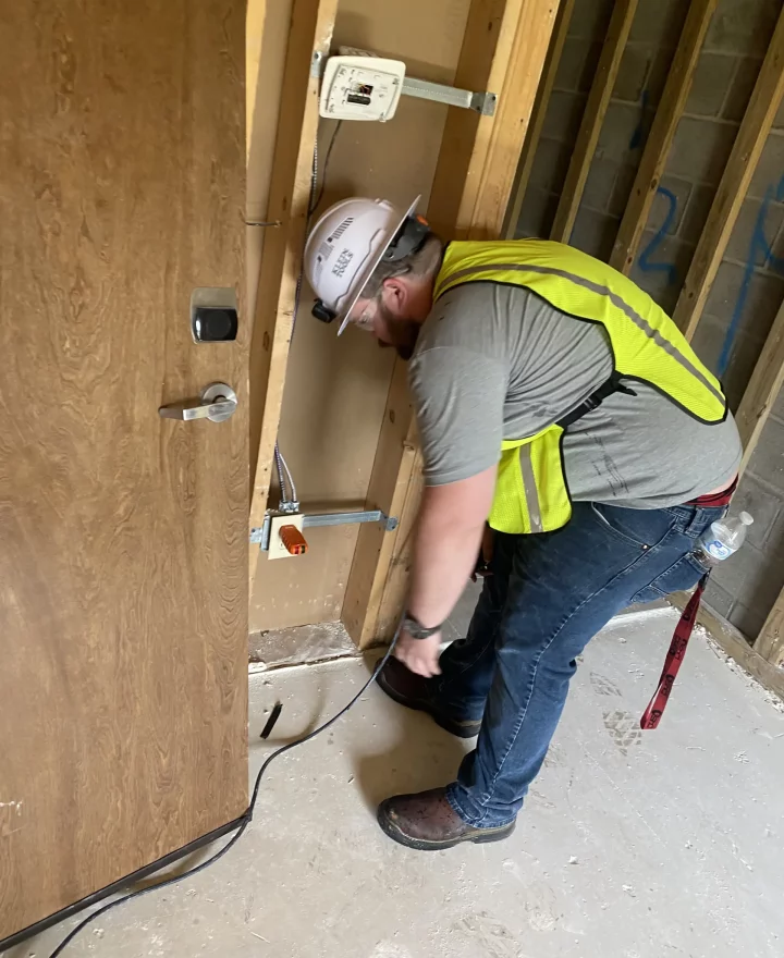 An electrician in a hard hat and reflective vest works on electrical wiring in a construction area.
