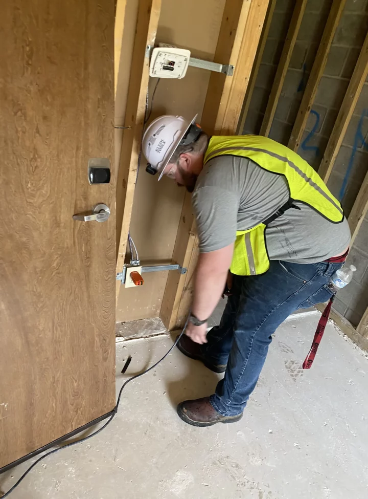 An electrician in a hard hat and reflective vest works on electrical wiring in a construction area.