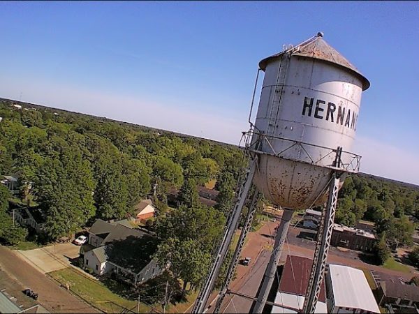 Aerial view of the weathered white Hernando water tower rising above tree-lined neighborhoods — a landmark symbolizing community roots and the need for resilient, local electrical infrastructure.