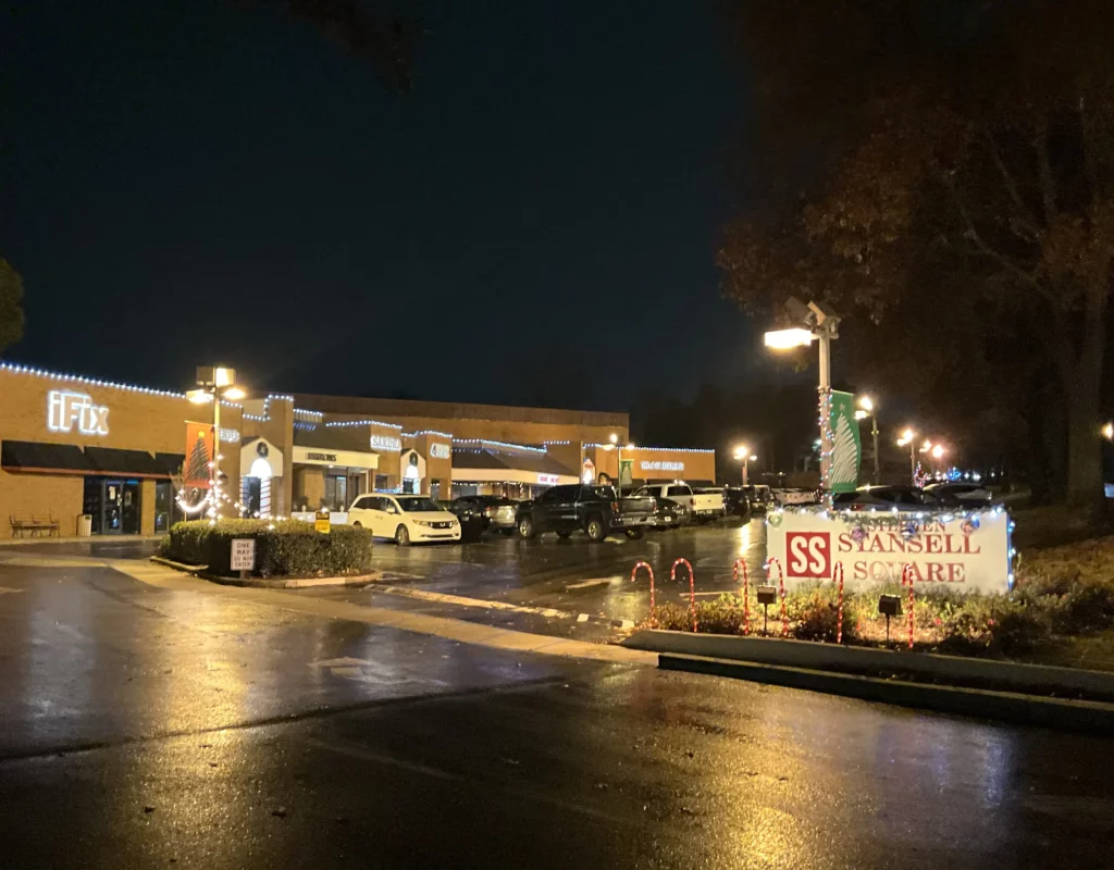 A nighttime view of a commercial plaza with festive lighting and decorations, showcasing businesses like 'eFix' and a holiday sign for 'Stansell Square.'