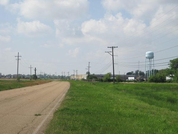 A quiet country road lined with utility poles and power lines stretching toward a distant water tower under a partly cloudy sky — symbolizing essential infrastructure in rural Mississippi.