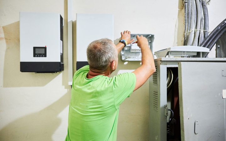 A technician in a green shirt working on an electrical panel with solar inverters mounted on the wall nearby.