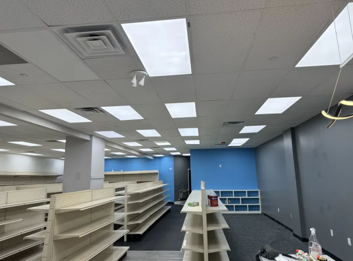 Wide-angle view of a commercial interior with exposed ceiling tiles, wiring, and store fixtures during renovation.