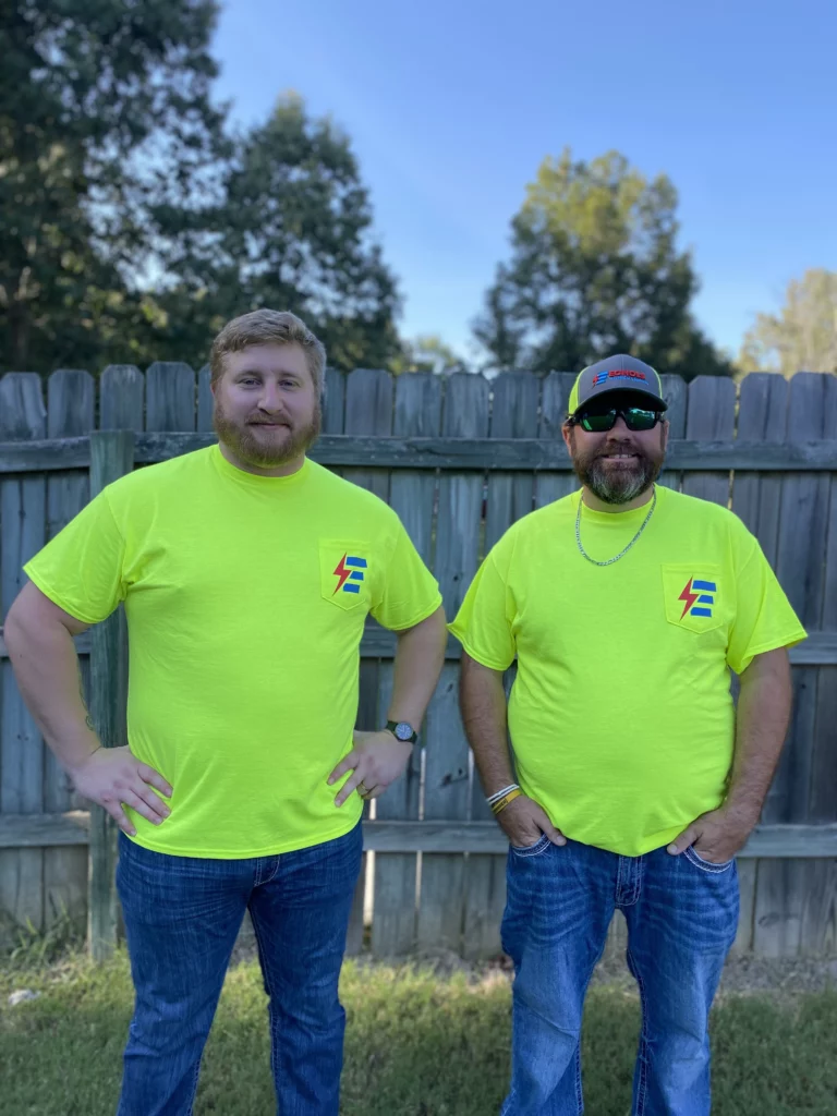 Two electrical service technicians in bright yellow shirts and jeans standing in front of a wooden fence.
